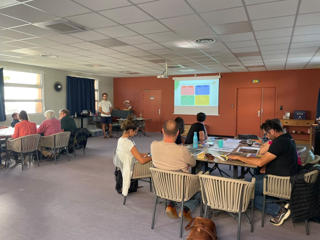 Workshop participants seated at tables in a conference room while a presenter stands near a projection screen displaying a slide. Attendees review printed materials and take part in a group discussion.