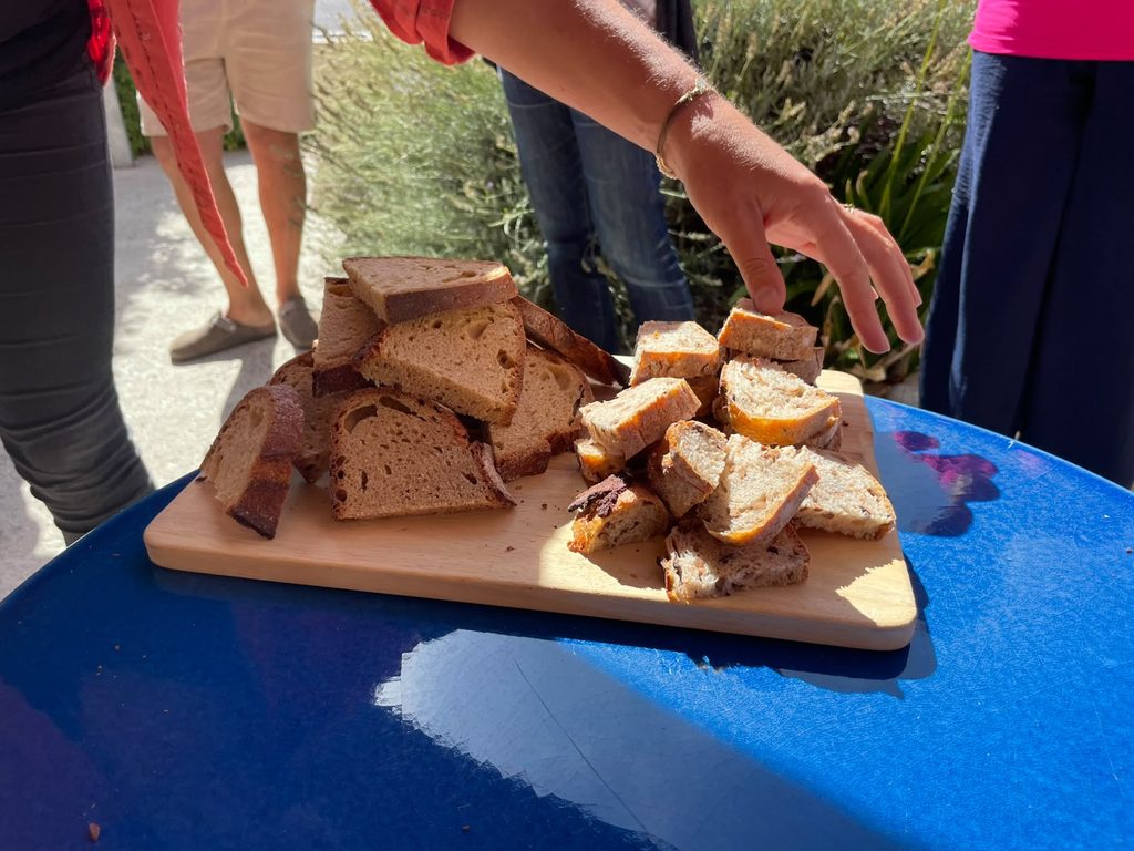 Close-up of sliced bread arranged on a wooden serving board placed on a blue outdoor table. Several workshop participants stand around the table, and one person is reaching for a piece of bread. The scene is brightly lit by sunlight, with garden plants and people visible in the background.