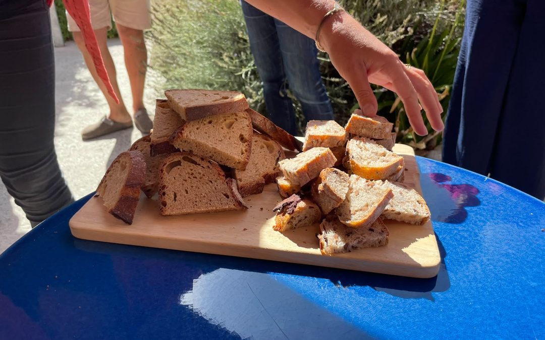 Close-up of sliced bread arranged on a wooden serving board placed on a blue outdoor table. Several workshop participants stand around the table, and one person is reaching for a piece of bread. The scene is brightly lit by sunlight, with garden plants and people visible in the background.