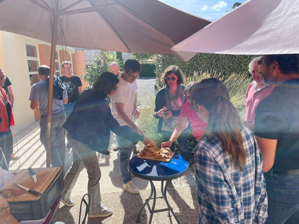 A group of workshop participants standing outdoors under large patio umbrellas, gathering around a small table to share pieces of bread during a break. Bright sunlight creates lens flare across the scene, with a building and garden visible in the background.