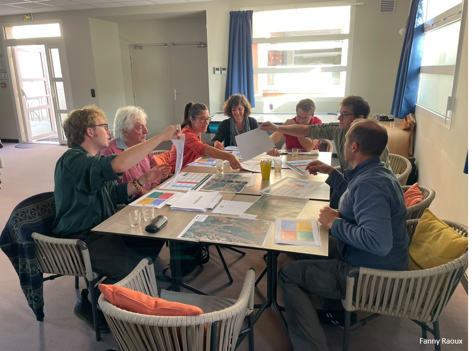 Participants seated around a meeting table covered with printed charts, maps, and documents, exchanging papers during a collaborative workshop session in a bright conference room.